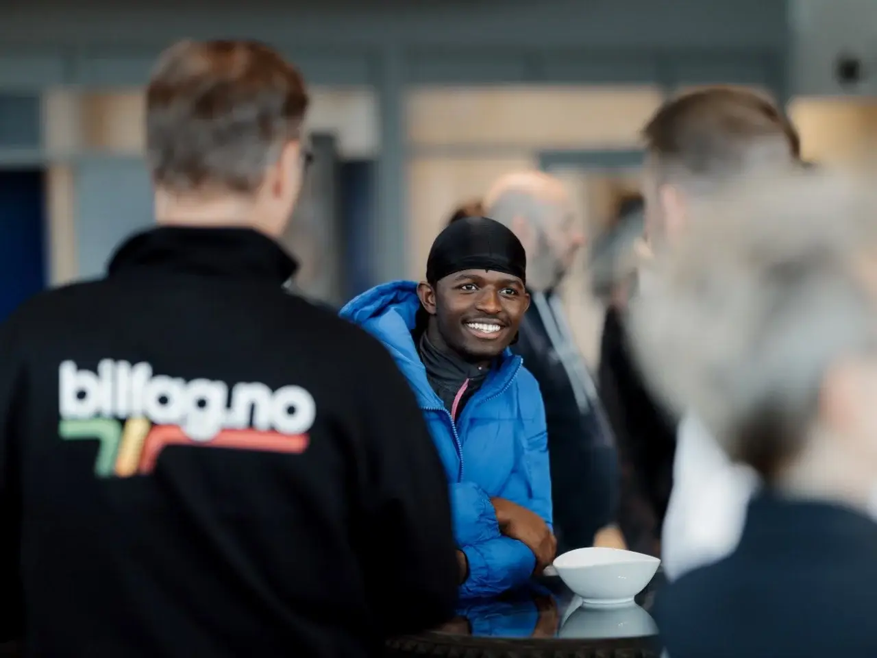 Smilende lærling i blå puffer-jakke og svart bandana ved kaffebord, samtale med bilfag-representant i forgrunnen
