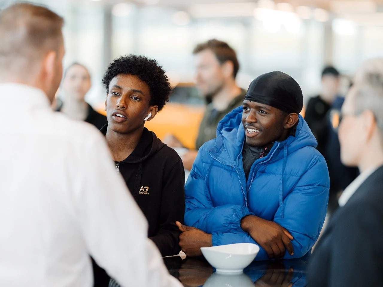 To smilende lærlinger ved kaffebord, en med AirPods i, samtale med kollega i hvitt skjorte i forgrunnen, orange Ford-bil i bakgrunnen