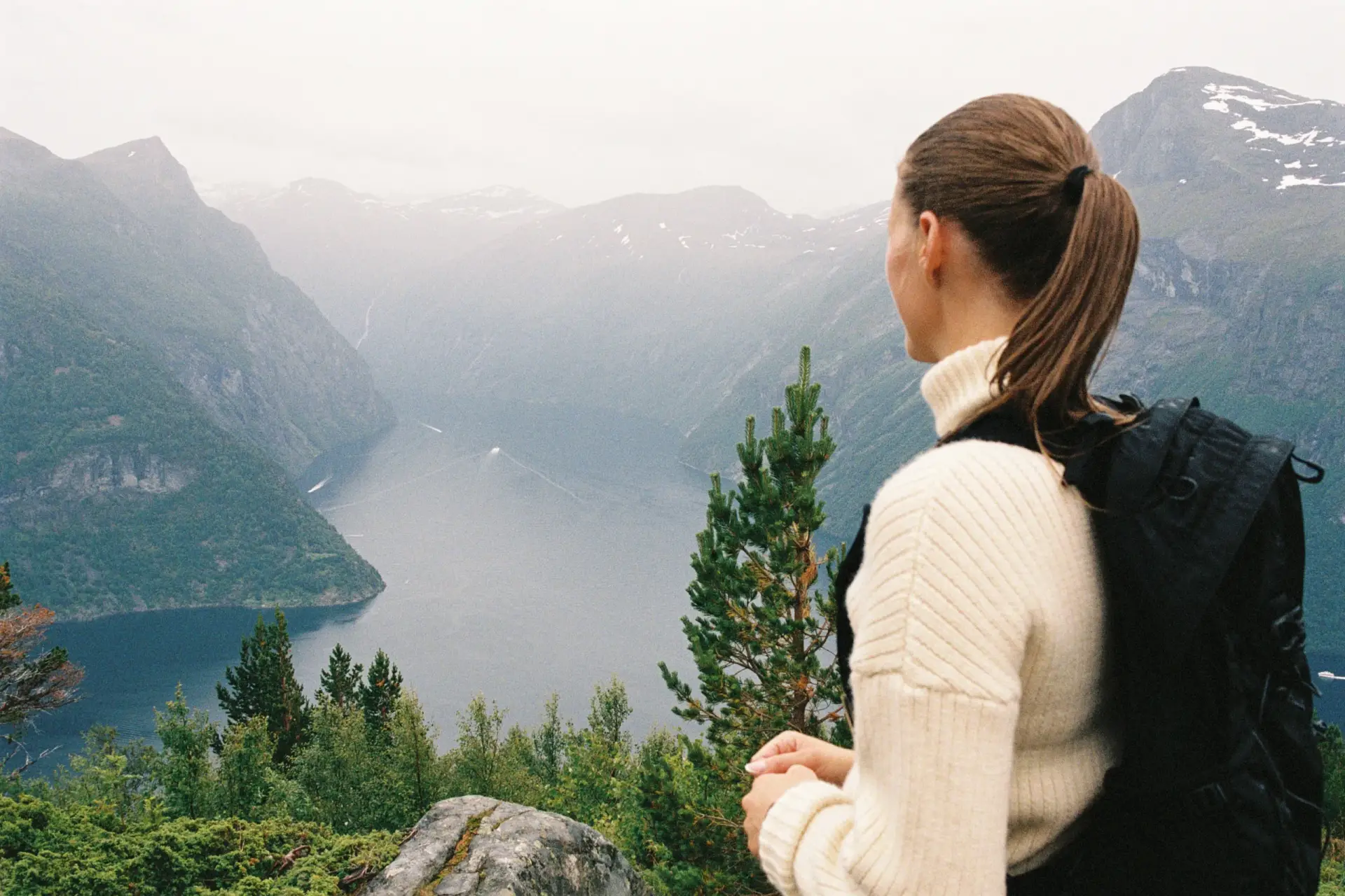 Kvinne i hvit ullgenser med ryggsekk ser ut over fjorden fra utsiktspunkt