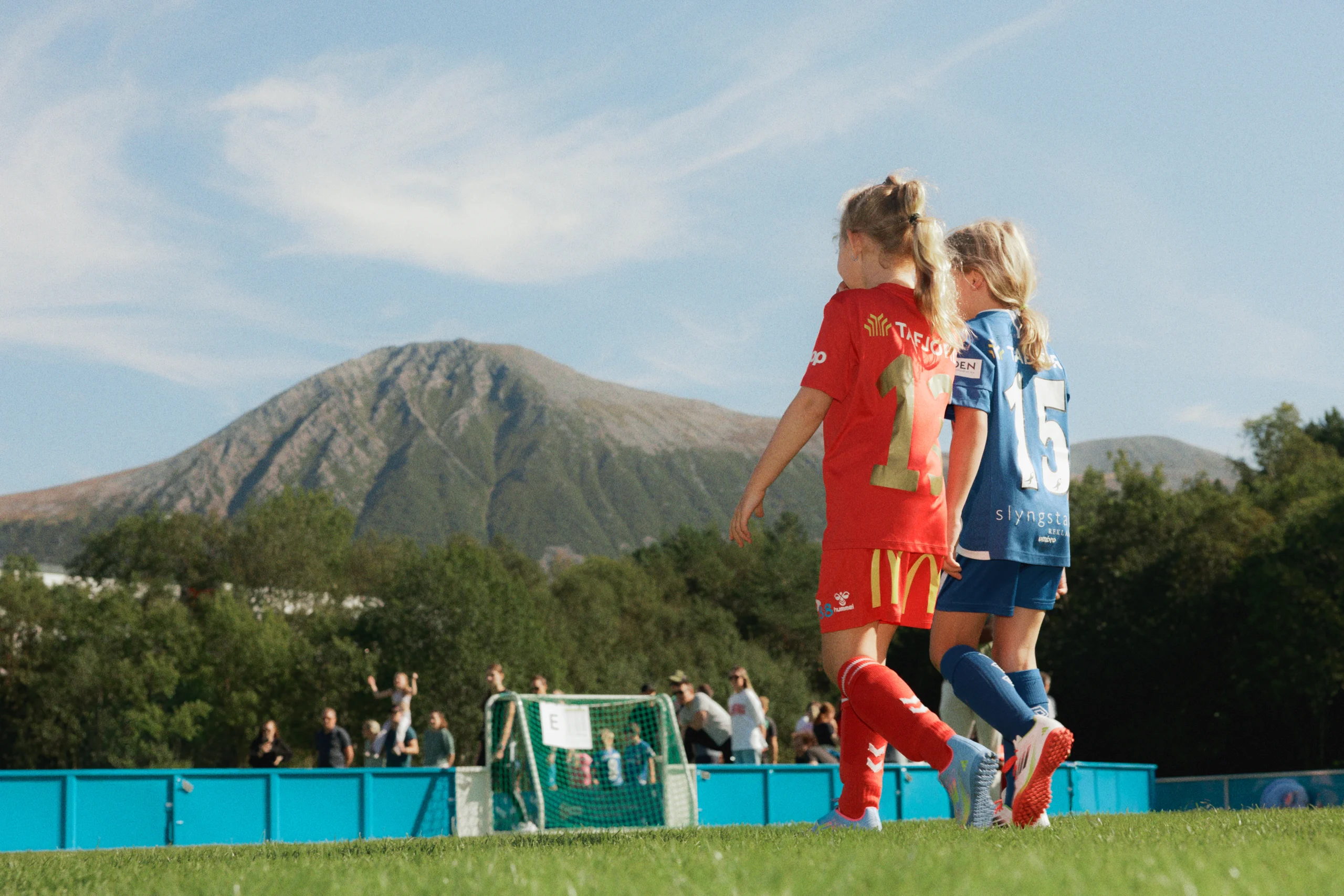 To unge fotballspillere — rød Spjelkavik IL og blå motstander — går side om side over banen sett bakfra, med stort Sunnmørs-fjell og varm sommerhimmel i bakgrunnen.