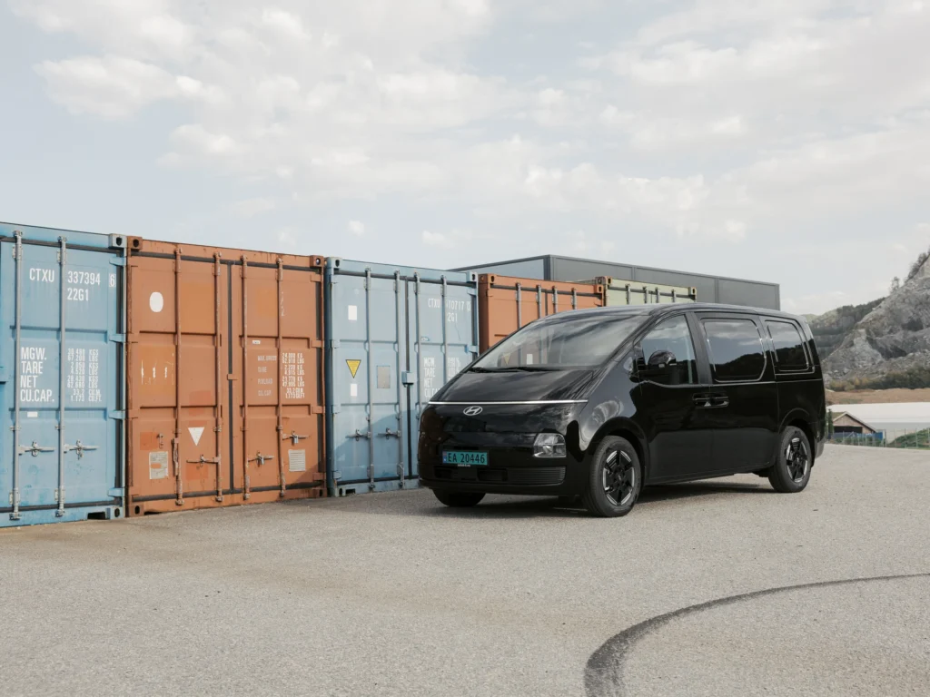 Svart Hyundai STARIA Electric varebil parkert foran en rad fraktcontainere